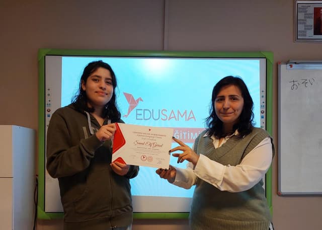 Student receiving a Japanese course certificate from her teacher in front of an Edusama slide